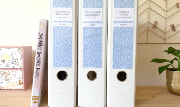 3 weiße Ordner mit Ordneretiketten von Alltagsfreuden in hellblau mit floralem Muster auf einem Sideboard
